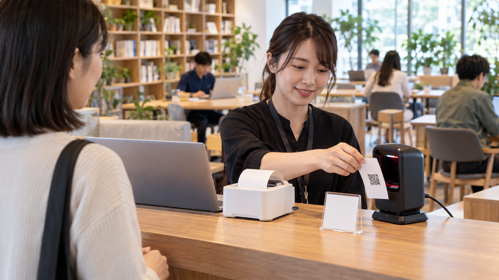 日本の明るいコワーキングスペースの受付カウンターで、スタッフが小型レシートプリンターから出たQRコード付き用紙を据え置き型QR対応バーコードリーダーにかざしている。隣に物理的な番号札とノートPCがあり、来場者が待っている。背景に書籍棚とフリーテーブル。実在企業のロゴ・社名なし、読める文字や個人情報なし、横長16対9の写真品質のイメージ。